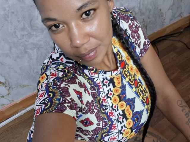 NaturaleBonyy with dark brown skin, braided hair, wearing a colorful, patterned top, looking up at the camera with a slight smile, showing a tattoo on her right arm.