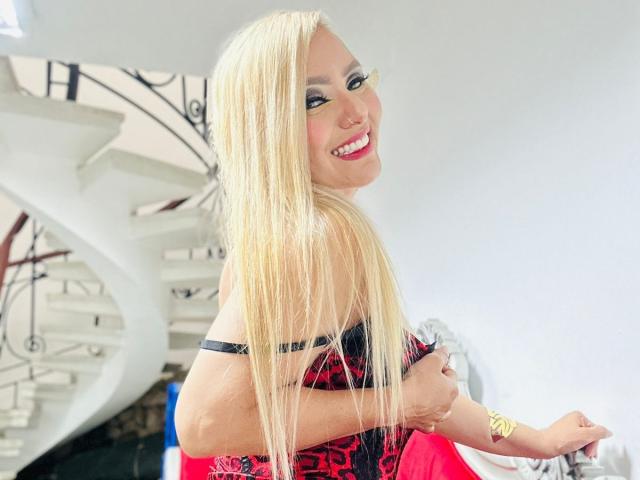 RoxannEmilss with long blonde hair, smiling, wearing a black and red lace corset, showing her tits, standing in a bright room with a white spiral staircase.