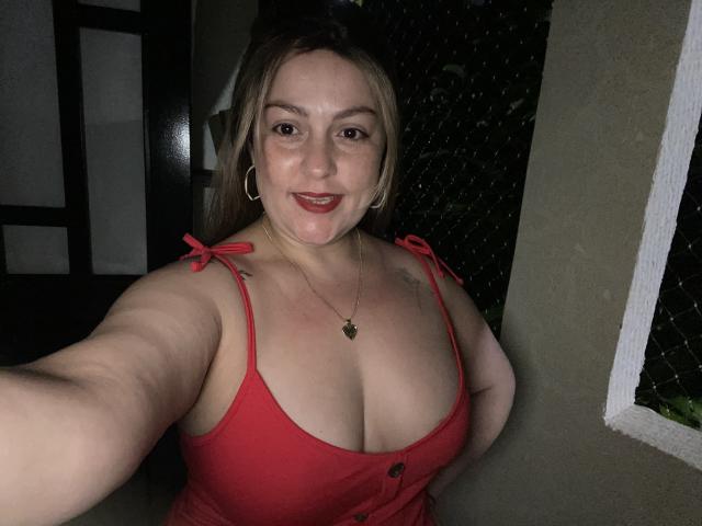 SamanthaGold with large tits in a red tank top, smiling selfie, showing cleavage, hoop earrings, and a necklace, in dim light, holding phone, background mostly dark.