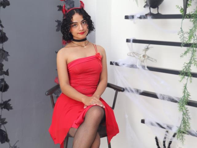 MiaWhalker with curly black hair, red devil horns, red strapless dress, black fishnet stockings, choker, sitting on chair, hands on knees, cute, red lipstick, smiling.