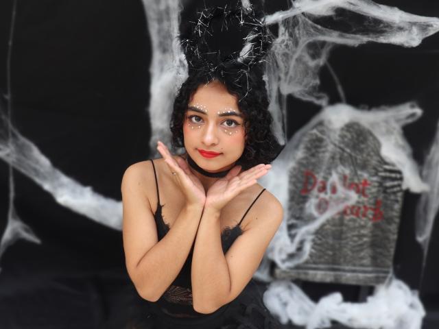 MiaWhalker with curly black hair, wearing a black dress, has white face paint and red lipstick, hands up, surrounded by smoke, black and white background.