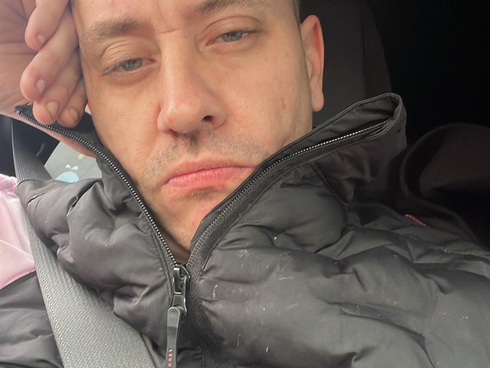 CalvinKyle with a serious face, wearing a black puffy jacket, slightly unzipped, with a seatbelt across his chest, hand on his head, looking tired and annoyed.