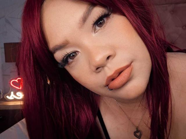 VictoriaField has red hair, brown eyes, and a nose piercing. She's wearing black lipstick and a necklace with a heart pendant. Her ass is not visible.