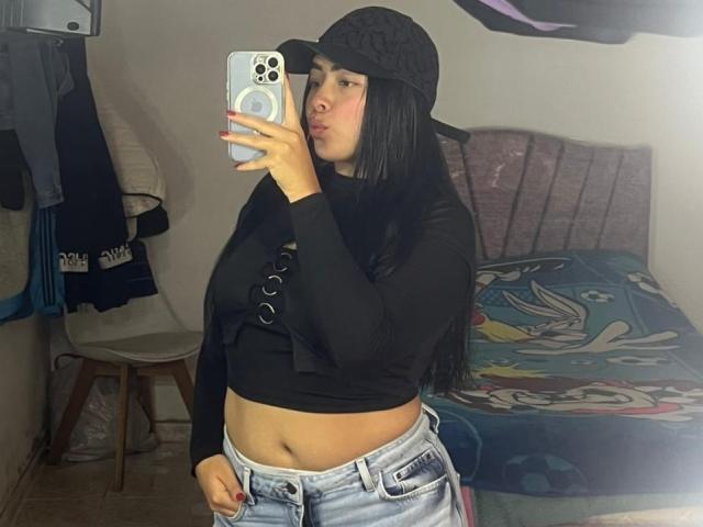 KateWelsin with long black hair, wearing a black cap, black long-sleeve top, and blue jeans, showing her bare midriff and slightly visible butt in a mirror selfie.