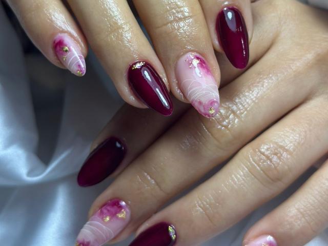 MiaDuboisX's hands show glossy, maroon-colored nails with pink and white gradient tips. Small gold dots are scattered on the pink tips. The maroon nails are glossy and smooth, contrasting with the translucent, gradient tips. The hands are placed over a white, silky fabric, highlighting the shine of the nails. The image focuses on the detailed, colorful nail art.