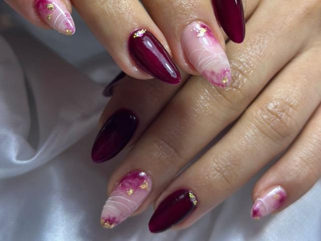 MiaDuboisX's hands with long, glossy nails. Most nails are deep maroon with a shiny finish, while the ring fingers are pink with white swirls and small gold accents. The pink nails have a semi-transparent look, blending with the maroon. Her fingers are slightly parted, showing the nails from multiple angles. The texture of the nails is smooth and reflective. The nails are well-manicured and polished. The skin tone is light. The image focuses on the nails and their detailed design.