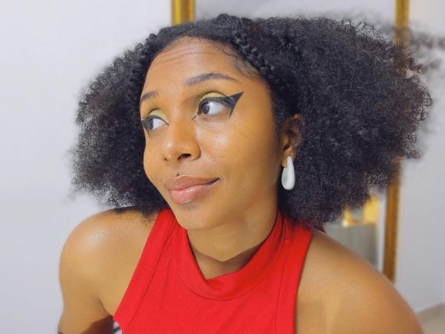 MeganWilliams with curly black hair, wearing a red sleeveless top, green eyeliner, black eyeliner wing, nose ring, and white teardrop earrings, looking to the side with a subtle smile.