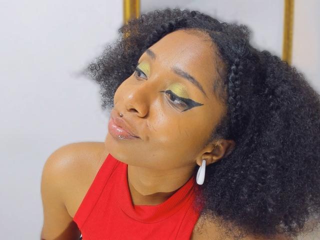 MeganWilliams has a dark, curly afro with a small braid on the left side. She wears a bright red sleeveless top and a white teardrop earring. Her yellow-green eyeshadow has bold black winged eyeliner. She has a small nose piercing and a shiny, slightly glossy lip. She looks downward with a slight, relaxed smile. The background is blurred and white, with a hint of gold in the top right. The image is clear and focused on MeganWilliams' face and upper torso.