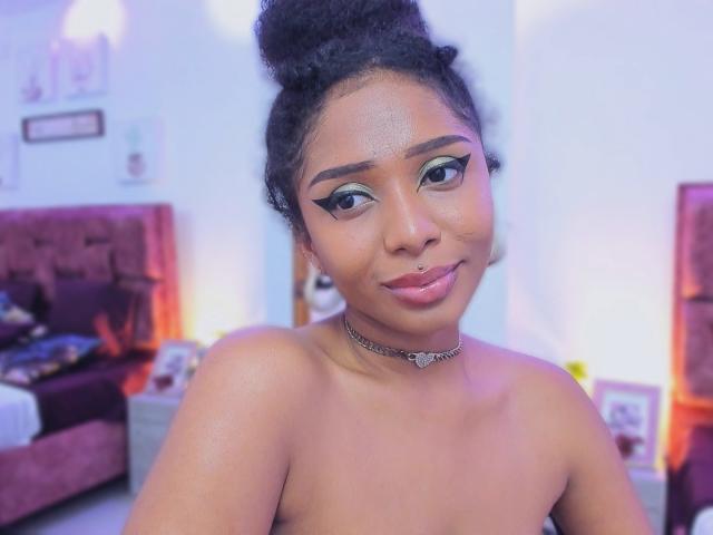 MeganWilliams, a dark-skinned woman with a curly updo, has shimmering skin and wears a silver choker. She has bold, green-blue eyeshadow, thick eyebrows, and a small nose ring. Her lips are glossy with a subtle pink tint. The focus is on her upper body, showing her bare shoulders. The background is blurred, featuring a pink bed and white walls. The image uses soft, warm lighting to highlight her face. She has a confident, slight smile.