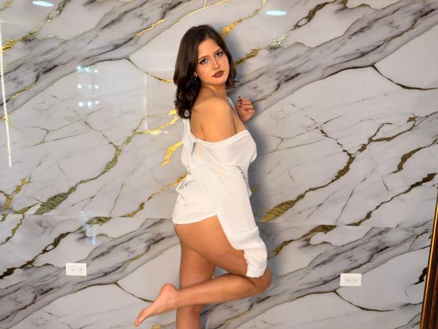 SofiaKennedy stands against a marble wall with grey and gold veining. She has dark brown hair, fair skin, and dark lipstick. She's wearing an off-shoulder white dress, exposing her right shoulder and upper back. She looks over her left shoulder, her right leg is bent, and her left leg is straight. Her right arm is slightly raised. The marble wall has two electrical outlets. The image has a watermark in the bottom right corner.