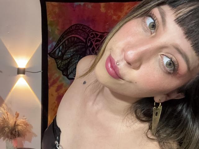 IzzysMisss with brown eyes, pink lips, nose piercing, and small tattoo near chin, wearing black top, gold earring, and black hair with bangs, in a brightly lit room.