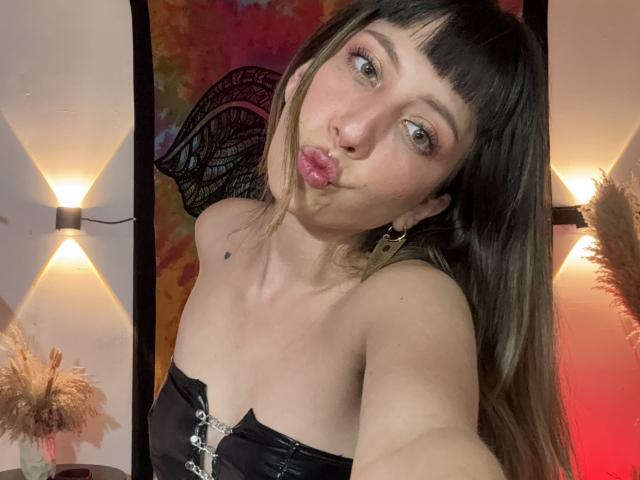 IzzysMisss with pouty lips, black strapless top, long brown hair, bangs, gold earrings, and a small tattoo on her neck, taking a selfie in a warmly lit room.