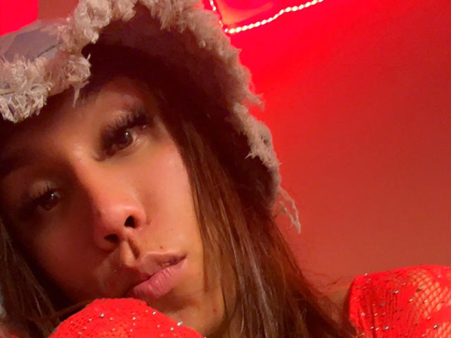 GeraldineSuan with brown eyes, wearing a fuzzy gray hat and red sequined top, pouting her lips slightly. Red light background. Boobs not visible, focus on face and hat.