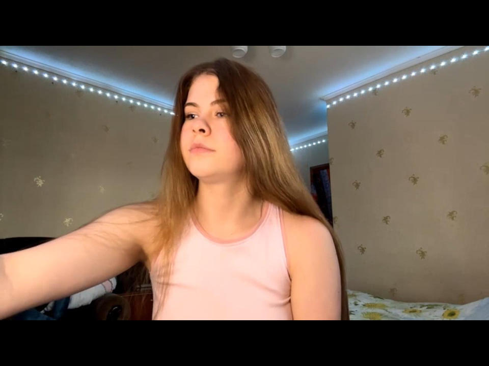 TennilleUsman with long brown hair, wearing a pink tank top, has a neutral expression, visible arm, and a subtle nose piercing, in a room with LED strip lights.