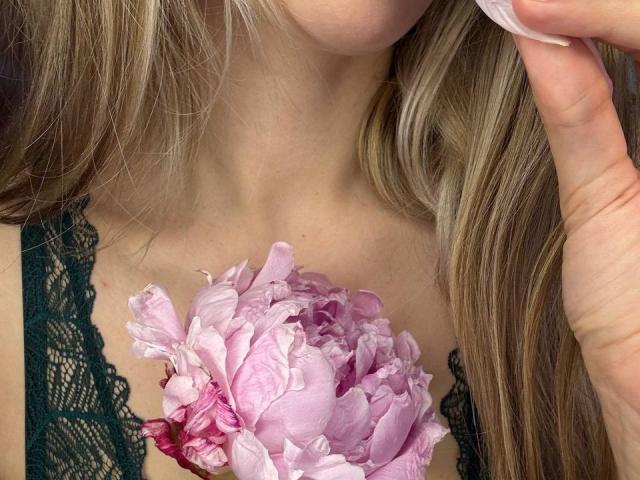 Close-up of MikyWayxy's upper chest and neck, with long, wavy blonde hair flowing to the right. MikyWayxy holds a pink, wet rose against their collarbone. They wear a dark green lace top. A hand with light skin and short, clean nails touches the top right corner of the image. The rose's petals are slightly open, showing a mix of pink and white hues. The image is detailed and realistic.