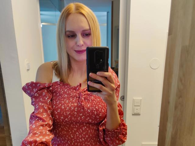 LaurenSommer-hot with blonde hair, pink lipstick, red floral dress, off-shoulder sleeves, holding black phone, taking mirror selfie, smiling slightly, white walls in background.