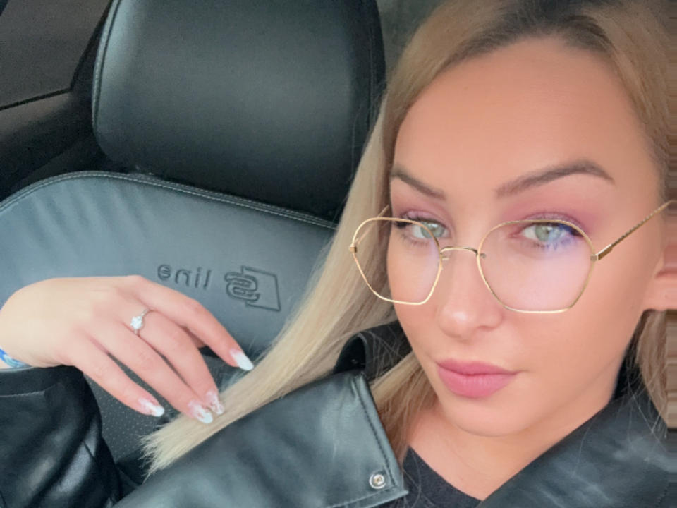 KristyKris with blonde hair, glasses, pink eyeshadow, and a ring on her finger, sitting in a black leather car seat, showing her nails and a slight smile.