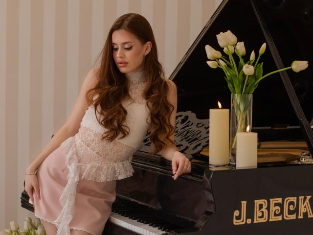 CherylFoxy stands next to a black J.Bechstein piano, leaning on it with her right arm. She has long, wavy brown hair and wears a white lace top with a pink, lace-trimmed skirt. Three white candles are on the piano, with a vase of white tulips in front of them. The piano has gold lettering on the lower right. The image has a soft, warm light. Her expression is calm, looking down slightly. The composition is elegant and refined.