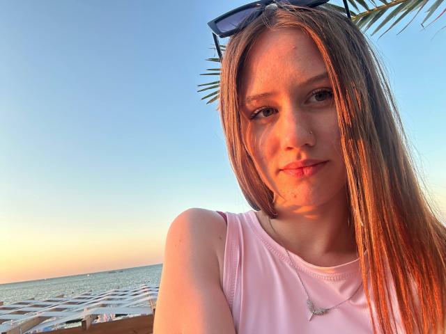 JamesZirkles with long brown hair, wearing a pink sleeveless top, blue sunglasses on head, slight smile, nose piercing, sunset light on face, palm leaves in background, ocean visible.