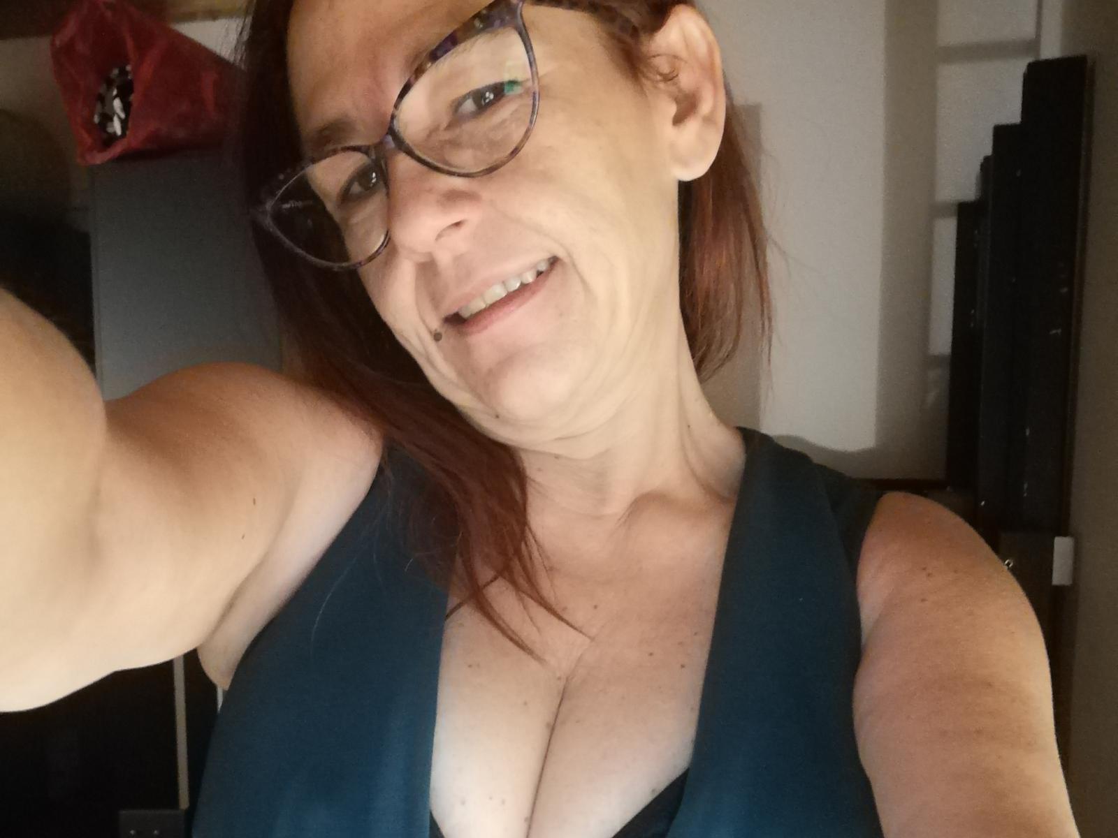 Klarissabloumm with glasses, smiling, wearing a green top that shows her big tits, has red hair, and is taking a selfie with her right arm.