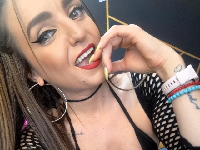 Close-up photo of WildBlondy6. She has light skin, dark brown hair, and is wearing bold, winged eyeliner with red lipstick. Her right hand, with yellow nail polish, is near her open mouth with a small yellow stick in it. She wears large silver hoop earrings, a black mesh top, and a black choker. Her left wrist has a white smartwatch, red bracelet, and blue bead bracelet. Tattoo visible on her right wrist. Background is blurred.