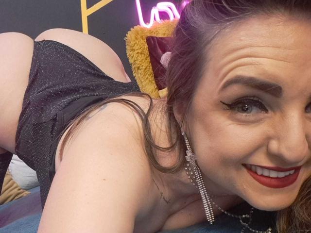 Close-up photo of WildBlondy6. She's on all fours, face close to the camera, smiling with red lipstick. Her brown hair is loose, and she's wearing sparkly black lingerie that reveals her buttocks. She has winged eyeliner, long eyelashes, and large earrings. Her skin is fair, and she's looking directly at the camera. Neon pink letters are in the background, and a yellow pillow is partially visible. The texture of the bed is soft and blue.