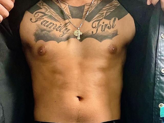 NigelBoydX's toned, bare chest with tattoos reading "Family First" above his nipples, wearing a black leather jacket, showing a cross necklace, and a flat, muscular stomach.