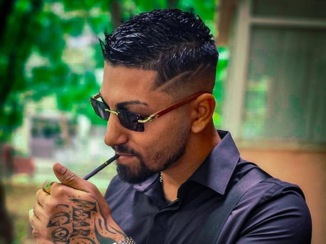 NigelBoydX with slick black hair, tattoos on right hand, dark sunglasses, black shirt, holding a cigar, looking down with a slight smile, standing outdoors.