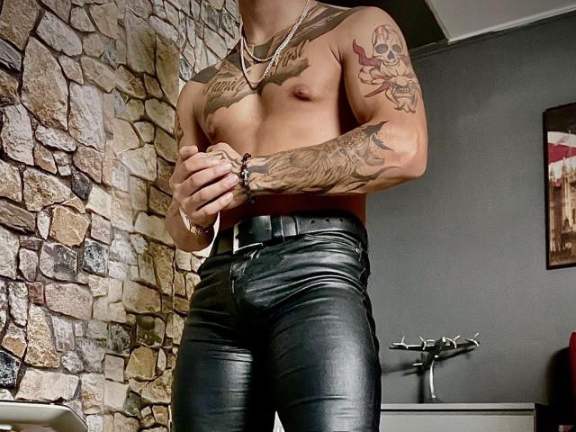 NigelBoydX, shirtless, with tattoos, wearing black leather pants, standing, hands in front, stone wall background, silver necklaces, muscular, tattooed arms, slightly visible chest and abs.