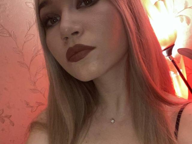 EssieVawter has long blonde hair, dark red lipstick, and a small necklace with a pendant. Her face is slightly angled with a subtle smile. Warm red light is in the background.