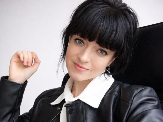 Liliit has short black hair with bangs, green eyes, and a light skin tone. She wears a black leather jacket with a white collared shirt. Her right hand is raised, fingers slightly curled. She has small gold earrings. The image is a close-up portrait with a simple, light background. Liliit's expression is calm, with a slight smile. The texture of the leather jacket is smooth, contrasting with the soft fabric of the shirt. Her hair is neatly styled.