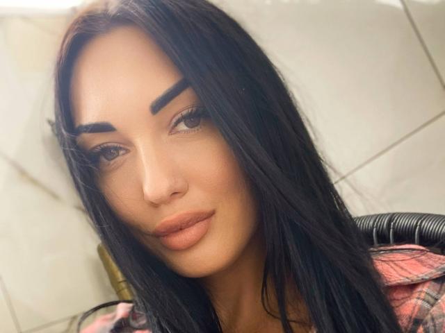 Xioni has long black hair, thick eyebrows, and full, slightly pouty lips. She's wearing a red plaid shirt, and her tan skin is smooth. Her eyes are half-open.