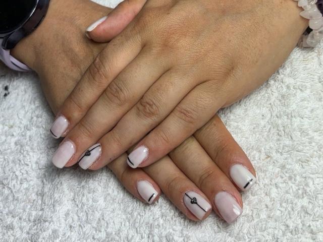 AmmaFrancaise's hands rest on a white textured surface. Her fingers are slightly spread, showing off a neat manicure. The nails are a soft pink with small black geometric designs on the pinky and ring fingers. Her skin is a warm brown tone. She wears a dark wristwatch on her left wrist and a white beaded bracelet on her right wrist. The nails are glossy and well-maintained. The composition is close-up, focusing on the hands and nails.