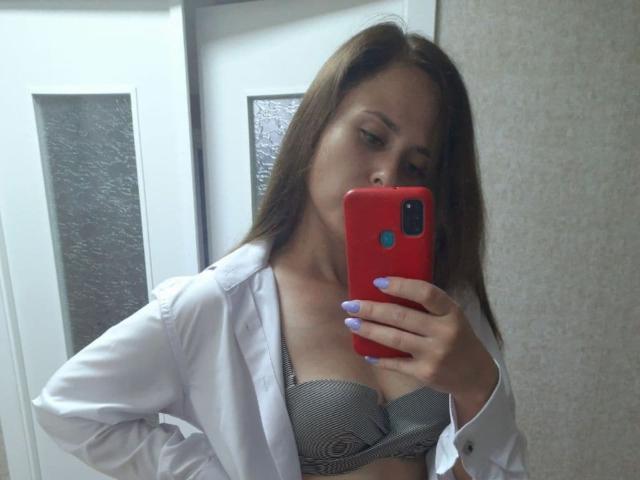 VictoriaAlarie taking a mirror selfie, wearing an open white shirt, grey bra with her tits visible, holding a red phone with purple nails, brown hair, and a neutral expression.