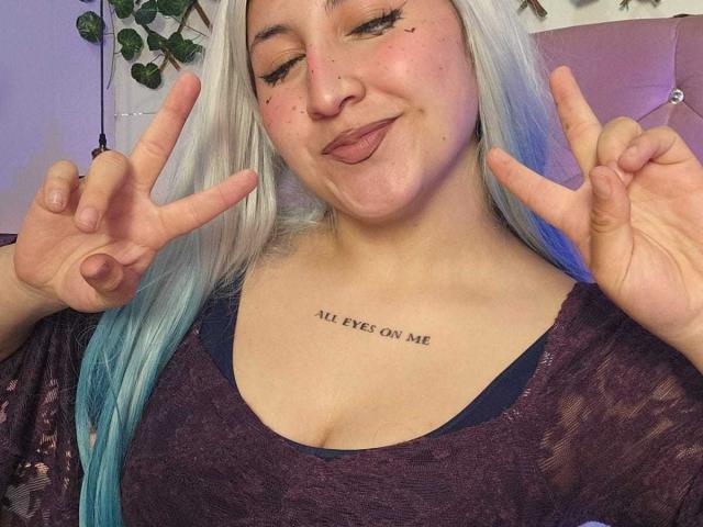 MaddieLesYoux with white-blue hair, making a double peace sign, wearing a purple lace top, and showing a chest tattoo. Her tits are small, and she's winking.