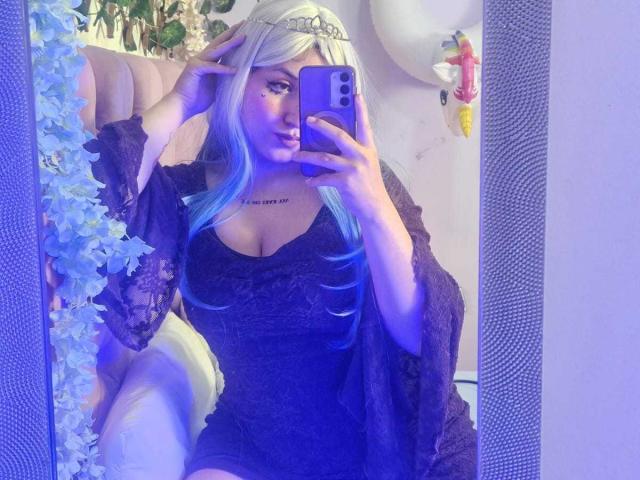 MaddieLesYoux takes a mirror selfie, holding a blue smartphone. She has long, blue-dyed hair, a silver tiara, and wears a black lace dress with a deep neckline. Her left hand touches her hair, and her right hand holds the phone. A black lace shawl drapes over her shoulders. She has a tattoo just above her cleavage. The blue and white reflection is highlighted with purple lighting. Her makeup is bold, with dark eyeliner and lipstick.