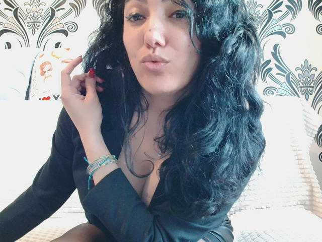 SexyMirra, a woman with curly black hair, sits on a white bed. She has light skin, a slightly pouty expression, and is wearing a black shirt with a deep neckline, revealing cleavage. She holds her hair with her right hand, showing red-painted nails. She wears blue bracelets on her left wrist. The background features black and white floral patterns. The image has a casual, slightly intimate feel. The overall composition is centered and focused on SexyMirra's face and upper body.