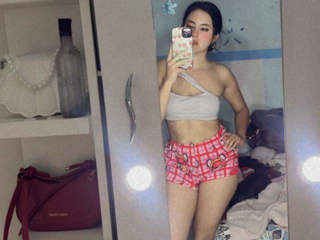 EimmyPark with small tits in a gray one-shoulder top and pink checkered shorts, standing in front of a mirror, holding a phone with a pink case.