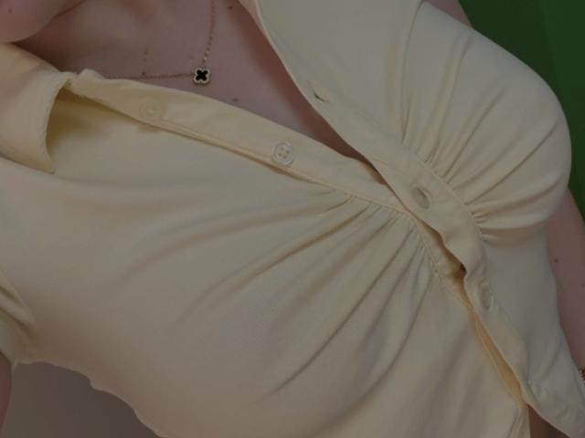Close-up of SugarrX's chest, showing a yellow button-up shirt with a gathered design, creating folds and a slight knot at the center. The shirt is slightly open, revealing a small portion of their skin. A delicate necklace with a small, black, cross-shaped pendant hangs around their neck. The shirt's fabric is smooth, and the color is a soft, pale yellow. The image focuses on the texture and form of the shirt and necklace.