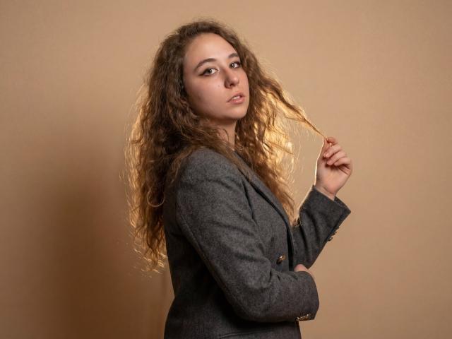 SaraJeredi with curly brown hair, wearing a gray blazer, holding a strand of hair, looking at the camera with a slight smile, brown eyes, soft makeup.