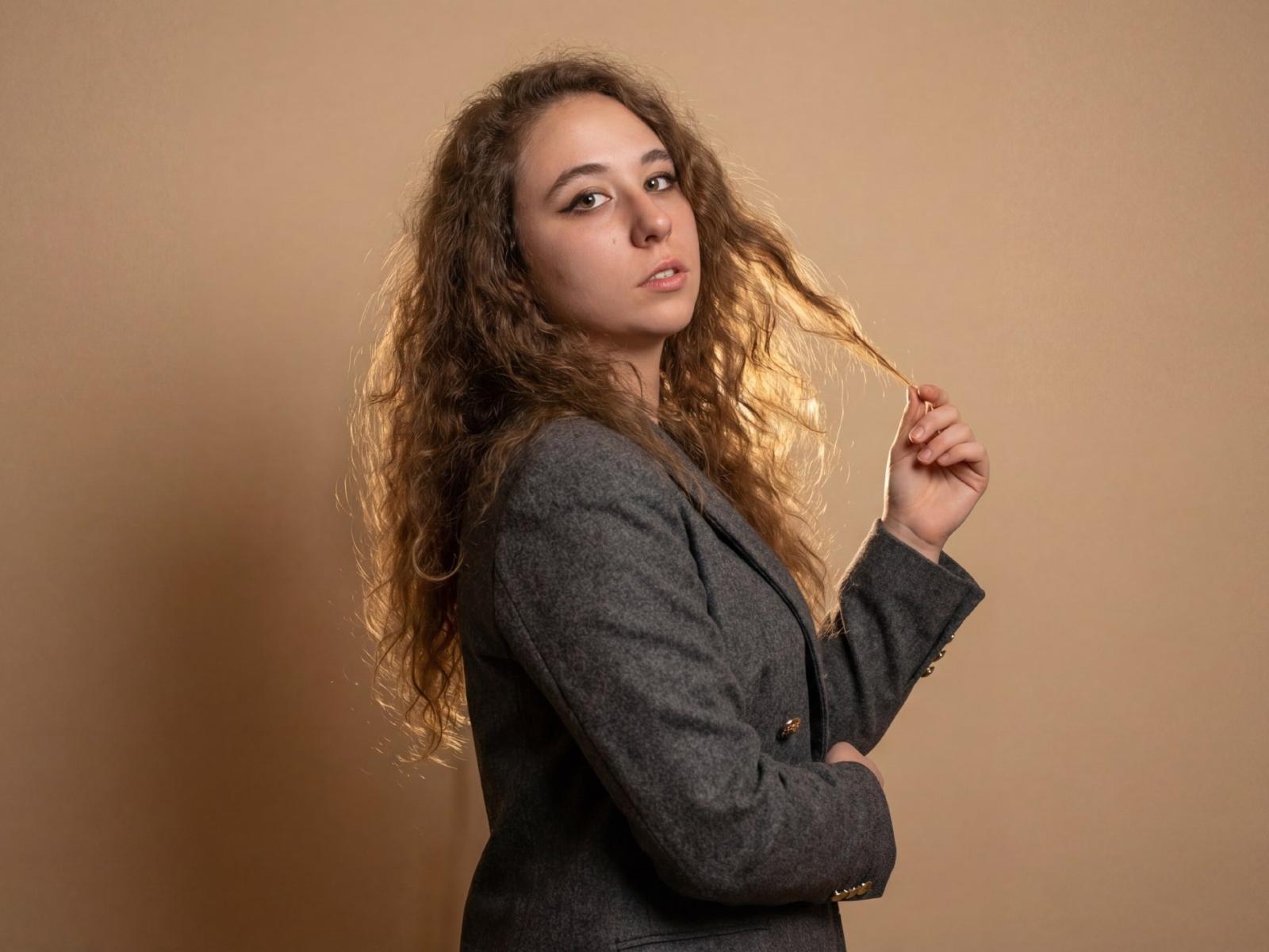 SaraJeredi with curly brown hair, wearing a gray blazer, holding a strand of hair, looking at the camera with a slight smile, brown eyes, soft makeup.