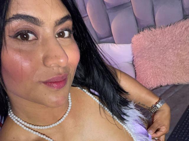 DiannaRosse has tan skin, black hair, brown eyes, pink lipstick, and a pearl necklace, with her hand on her white-feathered top, showing her tits. She's wearing a watch.