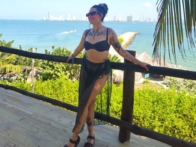 CarmilleZlata with tattoos, black bra, sheer green skirt, black sandals, standing on wooden deck, arms on railing, blue ocean, city skyline, palm leaves, smiling, dark sunglasses.
