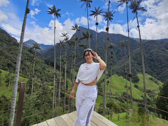 CarmilleZlata with dark hair, white crop top, and white pants with blue stripes, standing on wooden platform, palm trees, mountains, and green hills in background, touching her head.