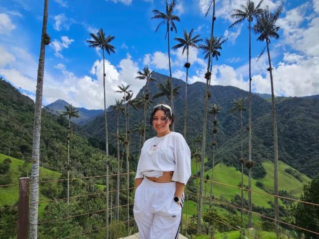 CarmilleZlata stands smiling, wearing a white crop top and high-waisted pants, hands in pockets, with palm trees and mountains in the bright blue sky behind her.