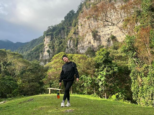 CarmilleZlata with a beard, black jacket, black pants, white sneakers, holding a red bottle, standing on a grassy hill, rocky cliff with greenery in the background.