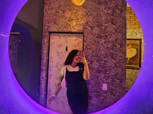 CarmilleZlata stands in a circular mirror frame with purple light, taking a selfie. She has long black hair, a tattoo on her right arm, and wears a black dress with a white collar. The background features floral-patterned wallpaper, a door with floral decals, and a framed painting. A warm light from above illuminates the scene. CarmilleZlata holds a phone with a colorful case in her right hand, smiling slightly. The mirror's circular shape and purple light create a vivid contrast.