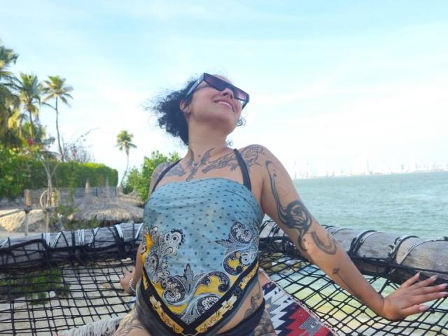 CarmilleZlata with curly black hair and sunglasses, leaning back on a netted float. She's wearing a blue, patterned strapless top and black shorts. Her arms and chest are covered in tattoos. She's smiling with her eyes closed. The float is in shallow, clear water with a palm tree and greenery in the background. The sky is bright blue with few clouds. The netted float has a black mesh pattern. CarmilleZlata's tattoos include floral and geometric designs. The image has a sunny, relaxed vibe.