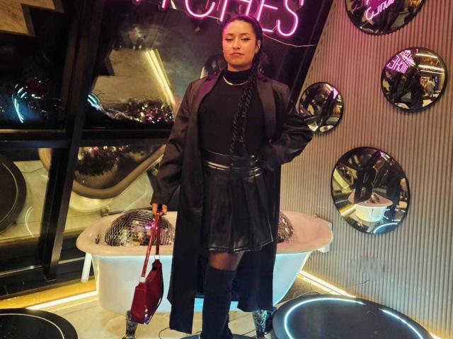 CarmilleZlata stands in a stylish interior, wearing a black turtleneck, black leather skirt, and long black coat. She holds a red handbag in her right hand. Her dark hair is in braids. The background features neon pink "CHICES" sign, round mirrors, and a white, vintage-style bathtub with a metallic dome. The room has a modern, slightly industrial feel with vertical striped wallpaper. CarmilleZlata's confident, slightly defiant posture is highlighted by her direct gaze.