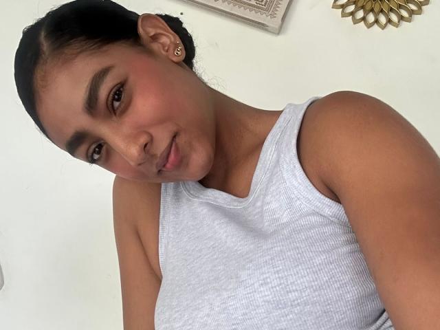 NatalyBlonder, a cute brown-skinned woman with dark hair in a bun, wearing a white tank top, showing her small tits and a subtle smile in a selfie.